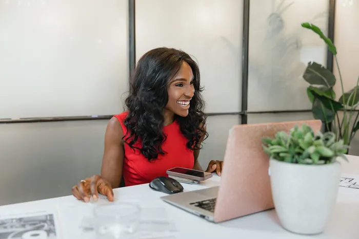 A woman in a red top smiles broadly at her desk in a bright office, laptop open in front of her.