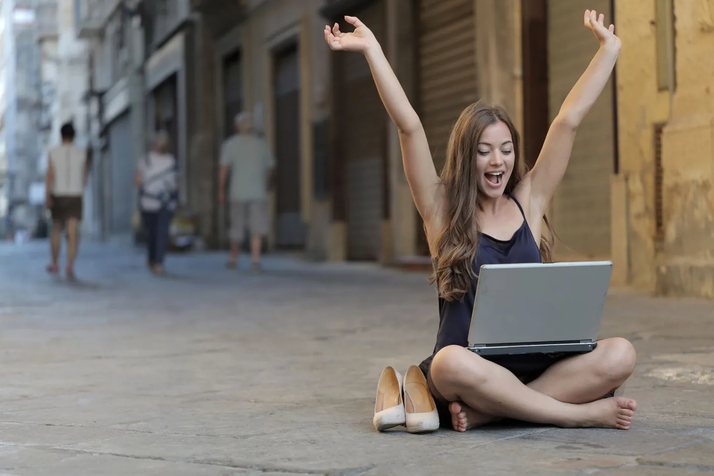 A woman sitting cross-legged on a city pavement throws both arms up in delight, a laptop on her lap.