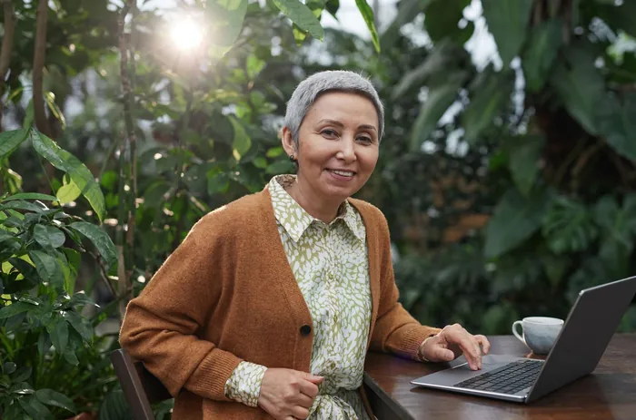 An older woman with short grey hair smiles from a table in a leafy garden, her laptop beside a cup.