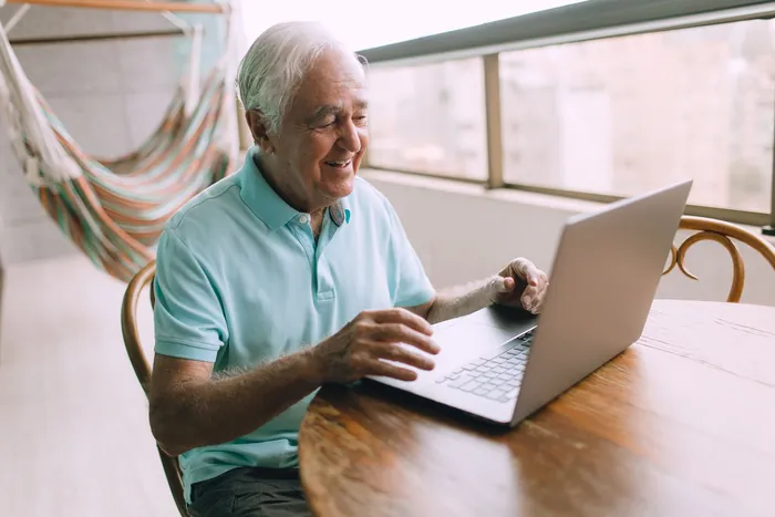 An older man with white hair smiles as he types on a laptop at a table by a sunny window.