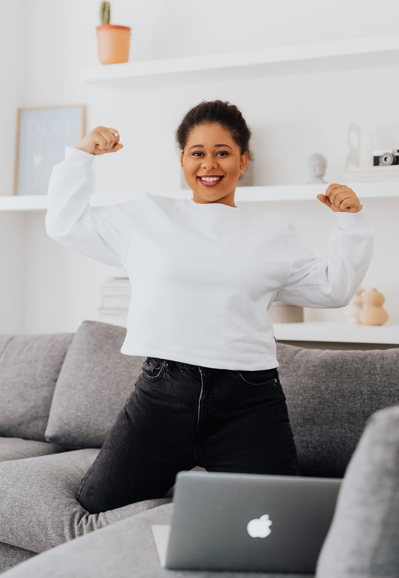 A young woman in a white jumper grins and flexes both arms in a bright living room, with her laptop on the sofa behind her.