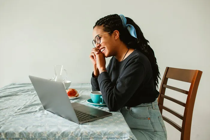 A woman with her hair tied back laughs at her laptop at a kitchen table, a cup beside her.
