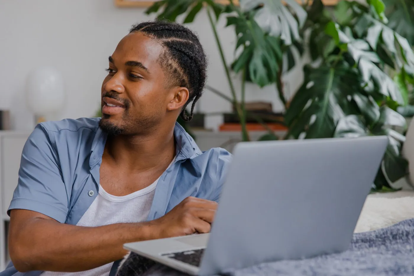 A man in a blue shirt sits at his laptop among houseplants, smiling as he looks away from the screen.