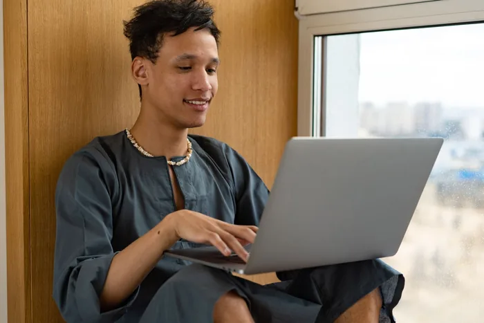 A young man sits in a window recess with a laptop on his knees, smiling as he types.